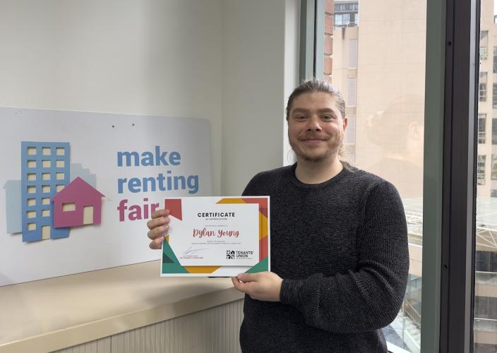 Dylan is smiling with his certificate. In the background there is a sign that says Make Renting Fair and a window looking over buildings.