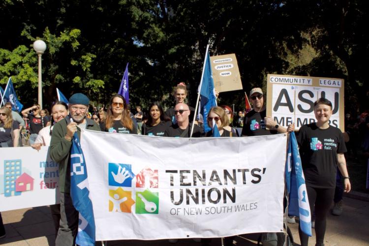 Tenants' Union members stand behind a banner with the Tenants' Union logo. People are smiling and wearing Make Renting Fair shirts with the purple and blue logo on them. There are trees and lots of people in the background.